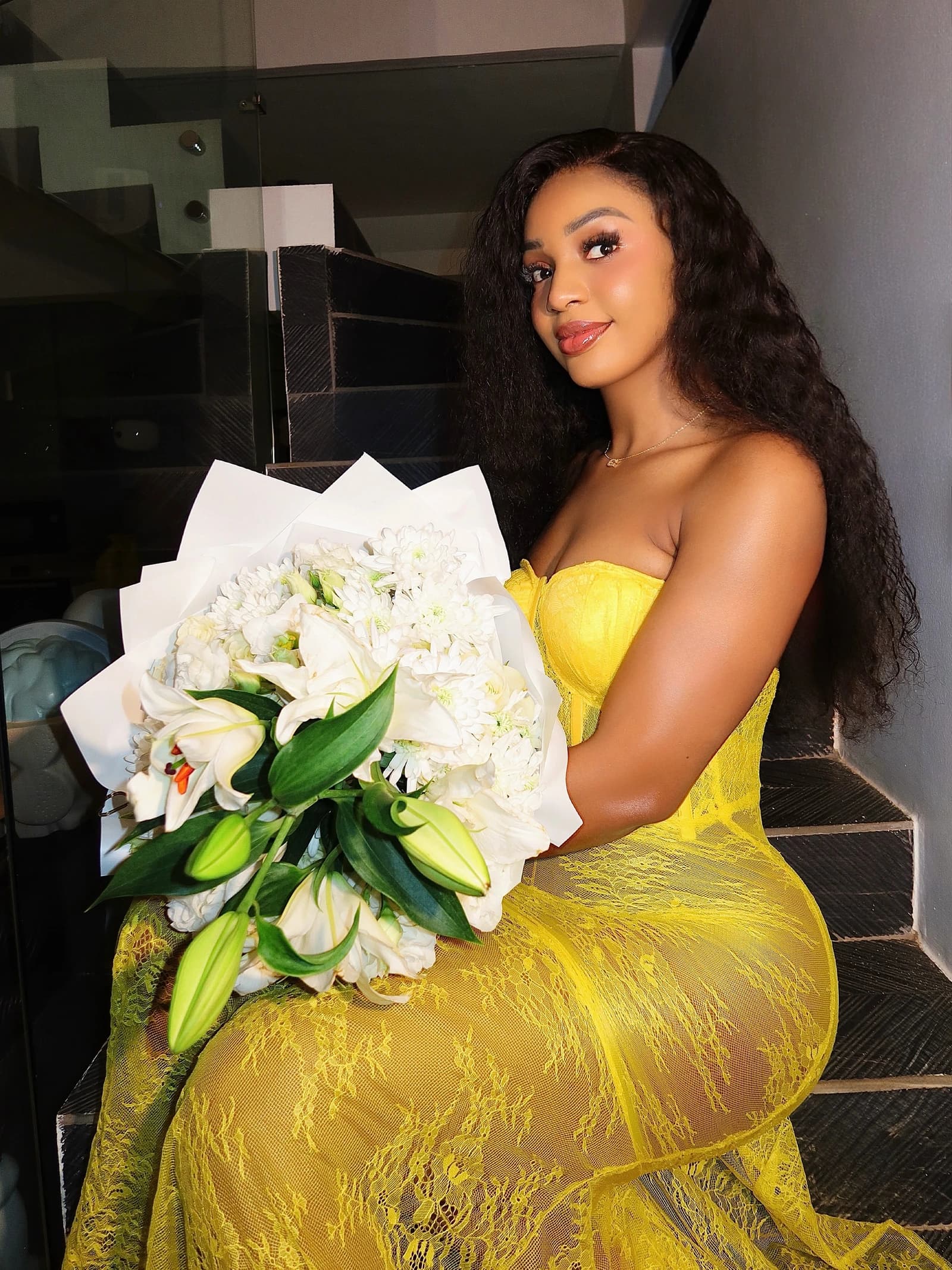 Portrait in a yellow lace dress holding a bouquet of white flowers on a dark staircase.